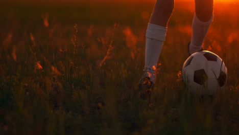 Close-up-of-Tracking-the-feet-of-a-football-boy-in-a-red-t-shirt-and-shorts-running-with-the-ball-at-sunset-in-the-field-on-the-grass.-The-young-football-player-dreams-of-a-professional-career-and-trains-in-the-field.-On-the-way-to-success